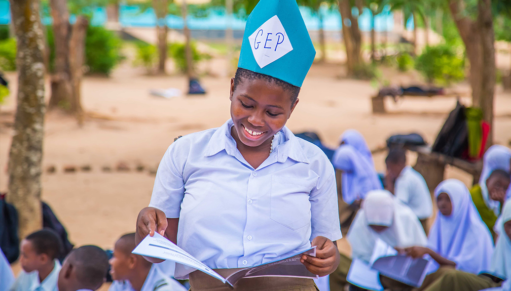 Girl from Tanzania standing at podium in front of her classmates wearing at hat that says "GEP"