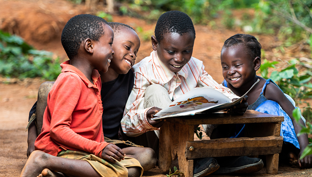 Group of young kids in Tanzania gathered outside around one big book reading and smiling