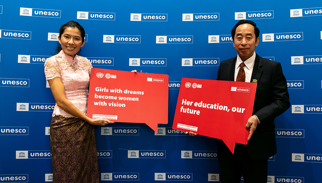 Two Cambodian staff members in front of UNESCO background holding signs promoting girls' education.