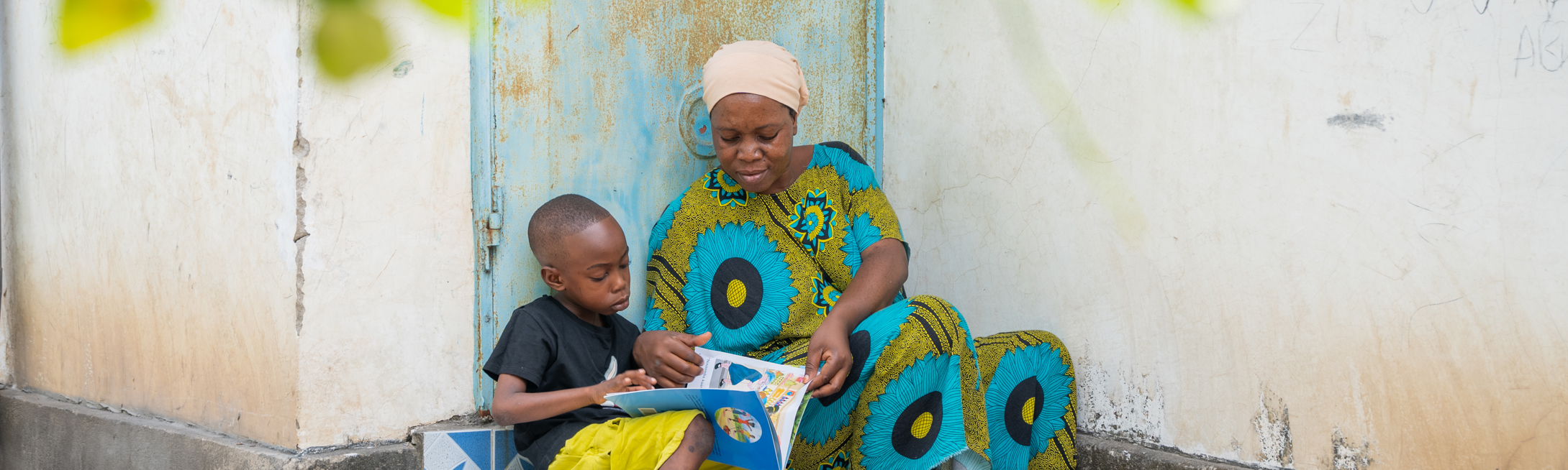 A Room to Read student in Tanzania reading with his parent on the steps of their home.